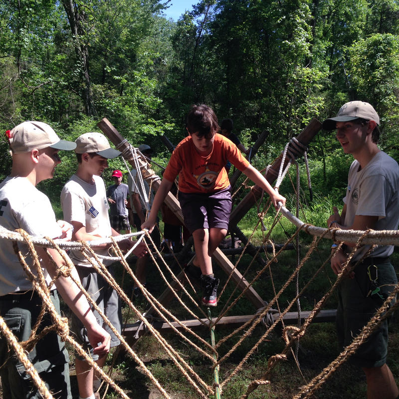 A boy walks down a rope bridge with staff members on either side