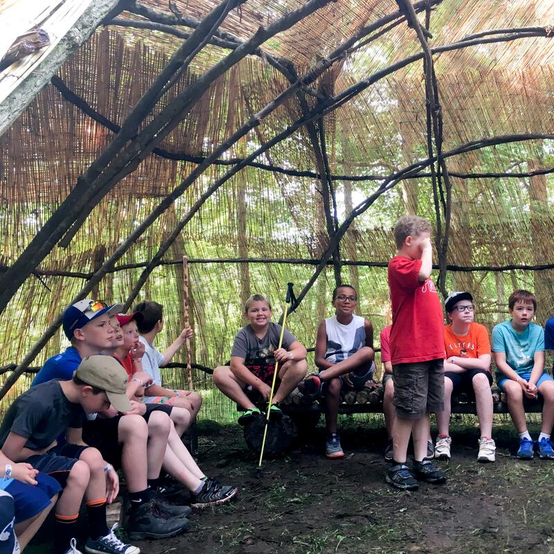 A dozen scouts gathered around inside of a wigwam made from natural materials