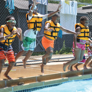 A small group of young campers in life preservers jumping into a pool